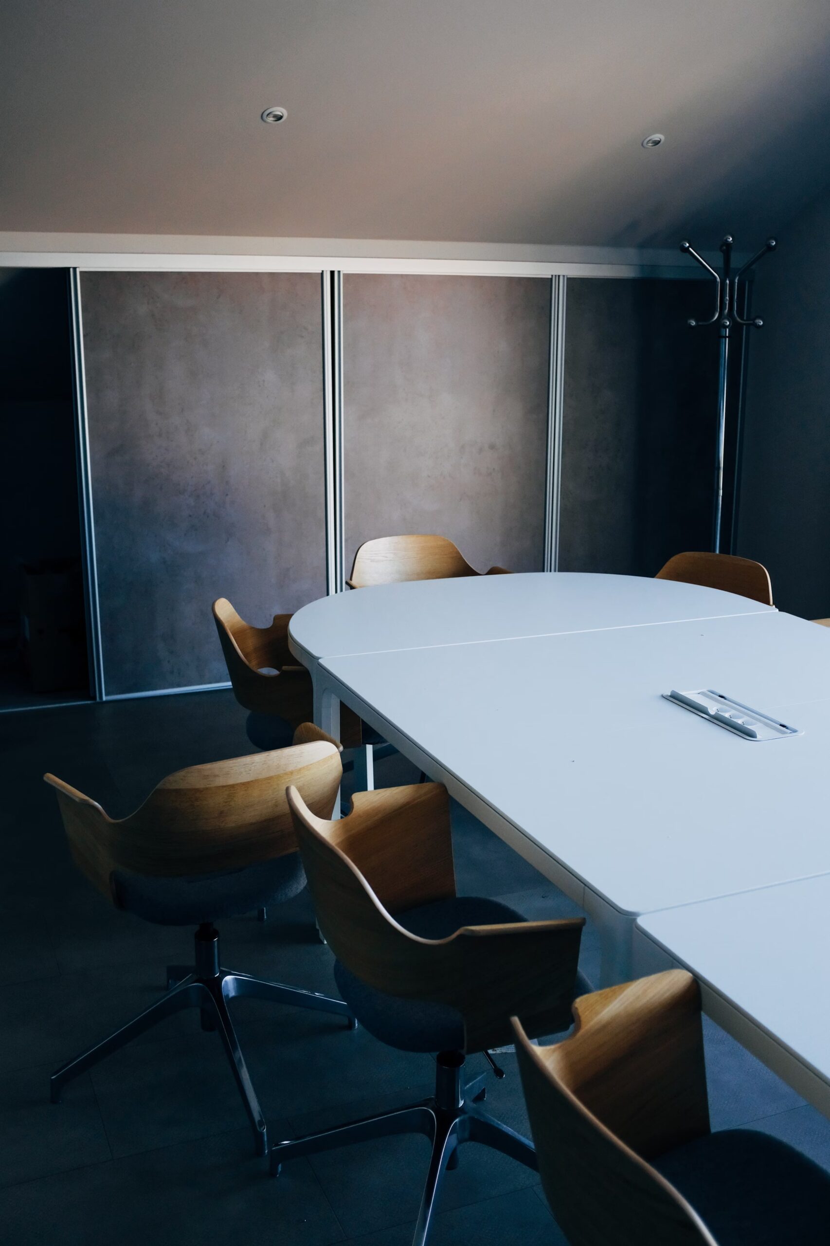 Sala riunioni minimalista con tavolo bianco e sedie in legno con base metallica, illuminata da luce soffusa.